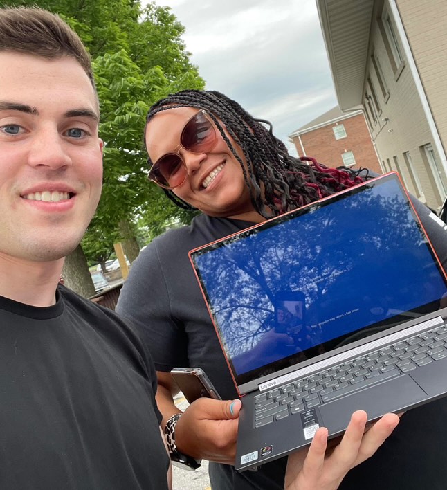 Two people outdoors holding a laptop. The laptop screen reflects trees and the sky. The person on the left is smiling, while the person on the right is holding the laptop and also smiling.