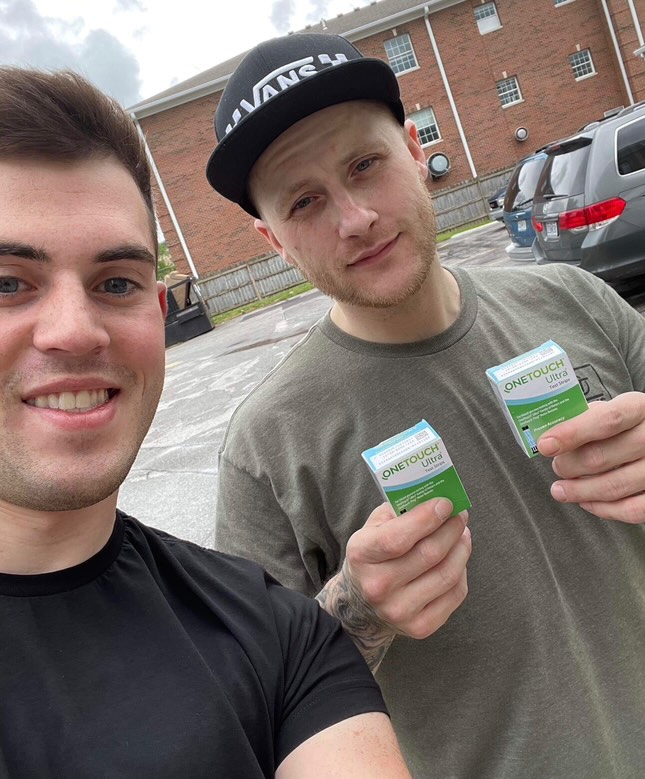 Two men standing outdoors. The man on the left is wearing a black shirt. The man on the right is holding two boxes labeled OneTouch Ultra, wearing a cap and a green shirt. Cars and a building are in the background.