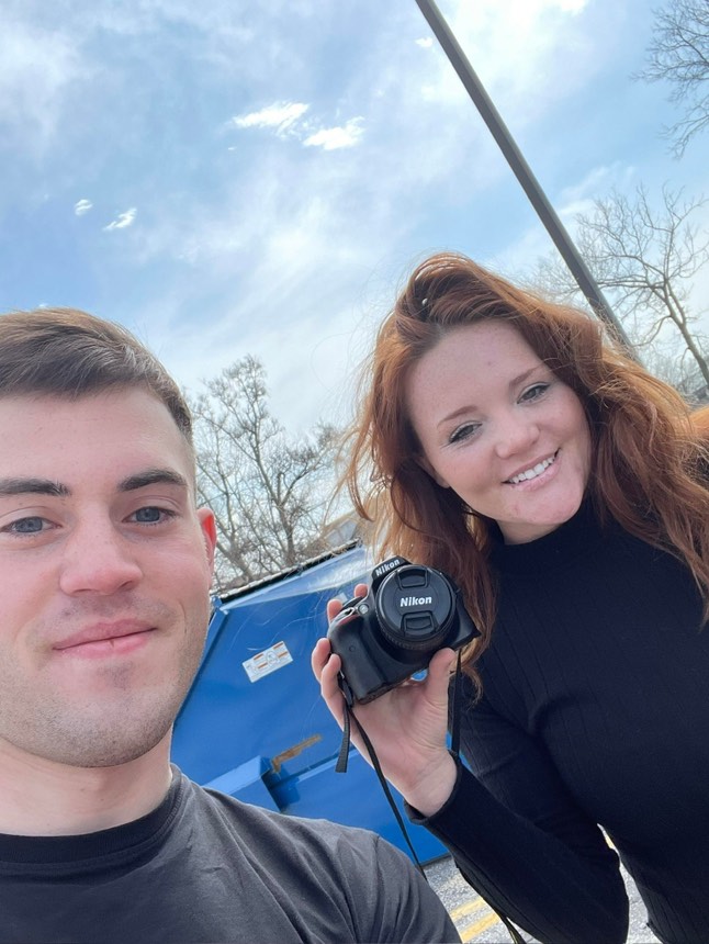 Two people smiling outdoors, one holding a Nikon camera. Blue dumpster and bare trees in the background.