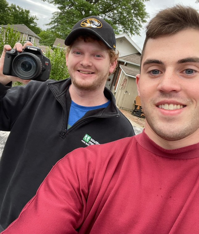 Two men taking a selfie outdoors. One man holds a Canon camera and wears a black cap and jacket. The other wears a maroon shirt. Background shows trees and a house.